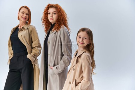 familia pelirroja feliz en elegantes abrigos posando con las manos en los bolsillos en gris, tres generaciones