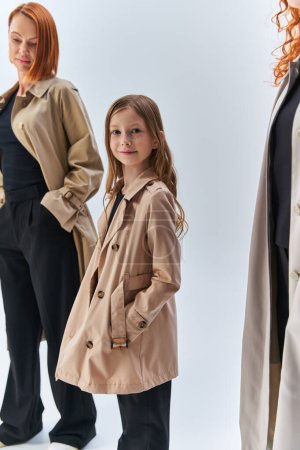 chica con abrigo posando con las manos en los bolsillos cerca de la familia pelirroja, tres generaciones de concepto de mujer