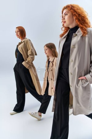 three generation redhead family walking with hands in pockets of trench coats on grey backdrop