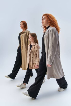 familia pelirroja de tres generaciones caminando con las manos en bolsillos de elegantes abrigos sobre fondo gris