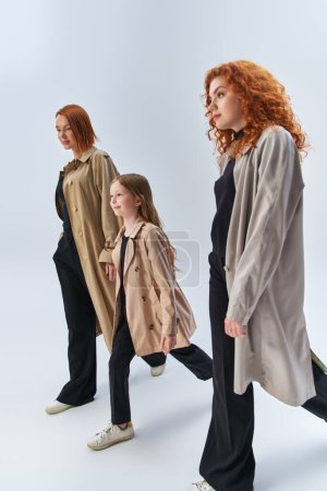 familia pelirroja de tres generaciones caminando juntos en elegantes abrigos sobre fondo gris, mujeres y niñas
