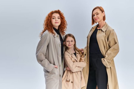 three generation joyful family standing together in stylish coats on grey backdrop, autumn fashion