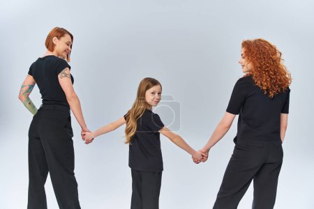 three generations, happy girl holding hands with women, standing in matching attire on grey backdrop