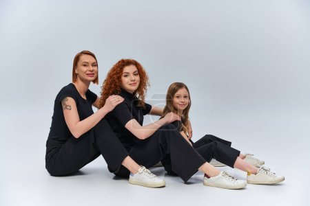 Drei Generationen Konzept, fröhliche rothaarige Familie sitzt in passenden Outfits vor grauem Hintergrund