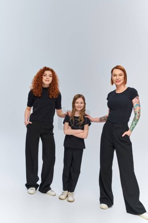 familia pelirroja en ropa a juego de pie juntos sobre fondo gris, tres generaciones femeninas