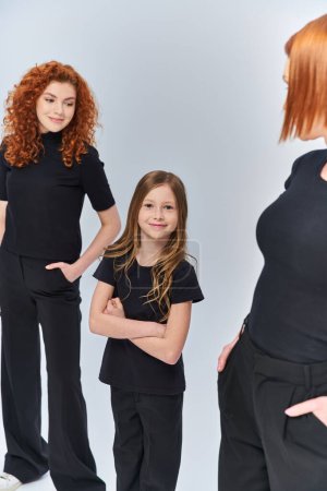 happy girl standing with folded arms near redhead family in matching clothes on grey backdrop