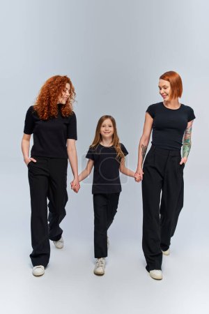 cheerful girl holding hands and walking with redhead family in matching outfits on grey backdrop