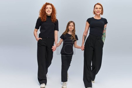 Photo for Cheerful girl holding hands and walking with redhead women in matching outfits on grey backdrop - Royalty Free Image