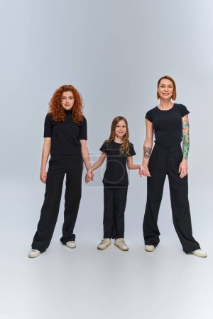 Photo for Cheerful girl holding hands and standing with redhead women in matching outfits on grey backdrop - Royalty Free Image