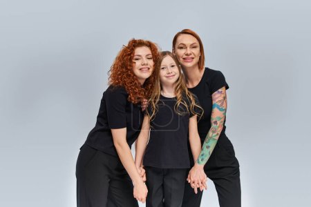 Photo for Positive girl holding hands and standing with redhead women in matching outfits on grey backdrop - Royalty Free Image