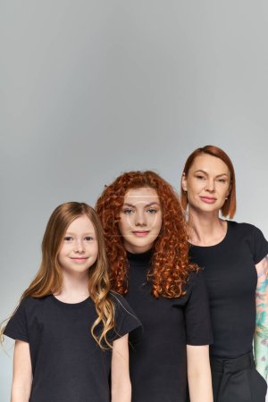 happy women red hair posing with girl in matching outfits on grey backdrop, three generations
