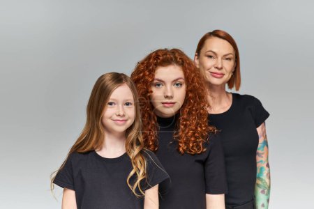 fröhliche Frauen mit roten Haaren posieren mit Mädchen in passenden Outfits vor grauem Hintergrund, drei Generationen