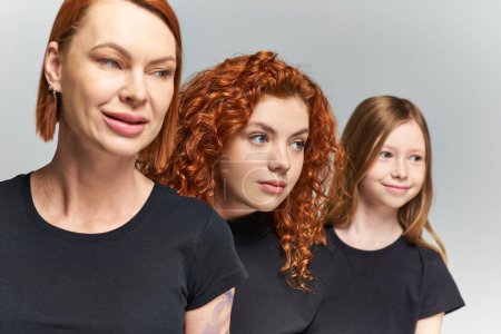 drei Generationen von rothaarigen Frauen und Mädchen posieren in passenden Outfits vor grauem Hintergrund, Familie