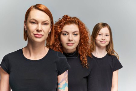 three generations of redhead women and child posing in matching attire on grey backdrop, family