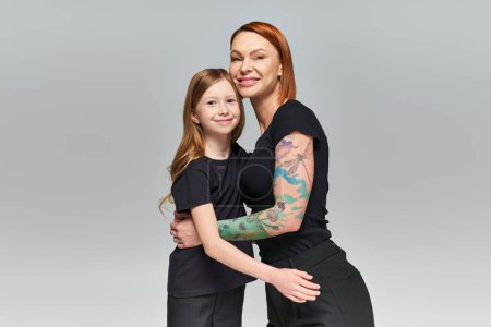 Photo for Two generations, redhead woman and girl in matching attire hugging on grey backdrop, joyous family - Royalty Free Image