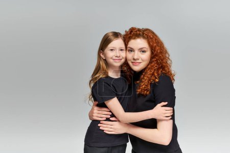 Zwei Generationen Konzept, glückliche rothaarige Mutter und Kind in passender Kleidung umarmen sich vor grauem Hintergrund