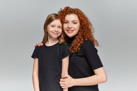Zwei Generationen Konzept, glückliche rothaarige Mutter und Tochter in passender Kleidung umarmen sich auf grau
