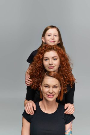 female generations concept, positive redhead family in matching attire hugging on grey background