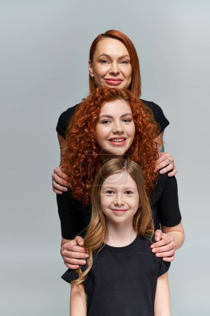concepto de generaciones, familia sonriente con el pelo rojo posando en trajes a juego sobre fondo gris