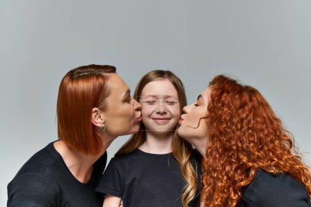 female generations concept, redhead women kissing smiling cheeks of girl on grey background
