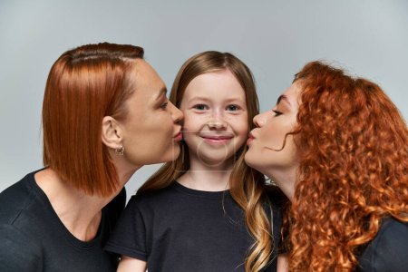 female generations concept, redhead women kissing cheeks of freckled girl on grey background
