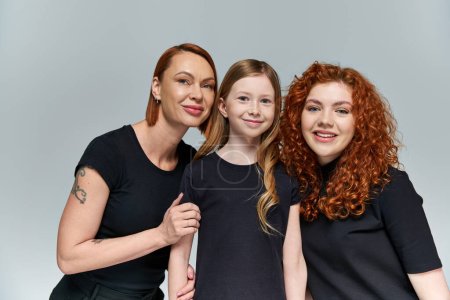 family bond, redhead women and freckled girl in matching attire smiling on grey background