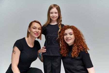 family portrait, happy freckled girl hugging redhead women in matching clothes on grey background