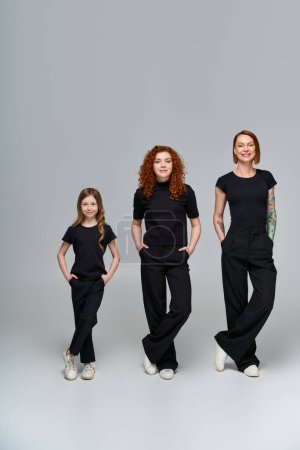 drei Generationen von Frauen posieren mit Händen in Taschen in passender Kleidung auf grauem Hintergrund