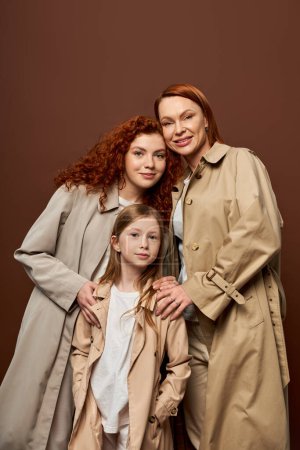 drei Generationen, fröhliche Frauen und Mädchen posieren in herbstlichen Trenchcoats auf grauem Hintergrund, Mode