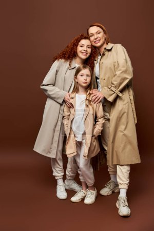 drei Generationen, glückliche rothaarige Familie posiert in herbstlichen Trenchcoats auf grauem Hintergrund, Mode