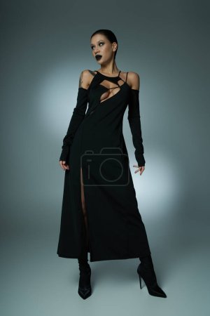 schauriger Glamour, verführerische Frau mit dunklen Lippen posiert in schwarzem Halloween-Kleid auf grau, voller Länge