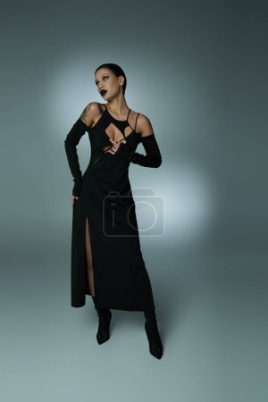 finstere Schönheit, Frau mit dunklem gruseligen Make-up posiert in schwarzem Halloween-Kleid auf grau, voller Länge