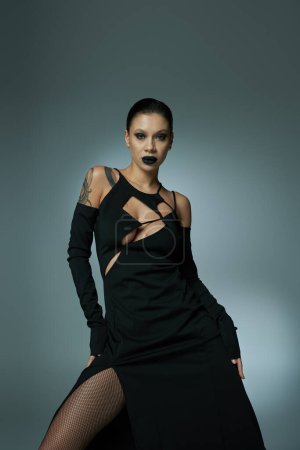 Schwüle Frau in schwarzem Kleid und dunklem, unheimlichem Make-up blickt in die Kamera auf grauem, halloween-artigen Konzept