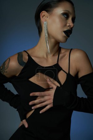 mujer tatuada en vestido elegante negro y maquillaje siniestro mirando hacia otro lado en el fondo azul y gris
