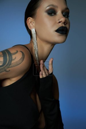 retrato de mujer encantadora tatuada con maquillaje oscuro mirando a la cámara en el fondo azul y gris