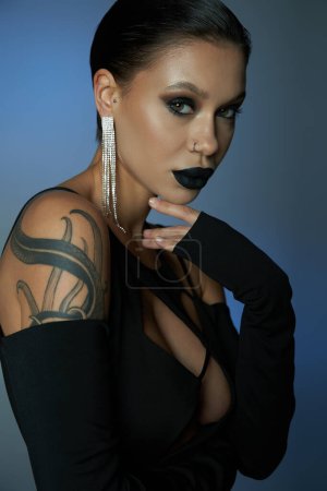 tattooed woman with dark eerie makeup looking at camera on blue and grey backdrop, halloween concept