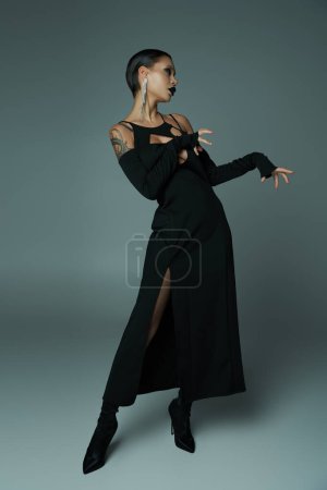 dark beauty, tattooed woman in black halloween dress and eerie makeup posing on grey, full length