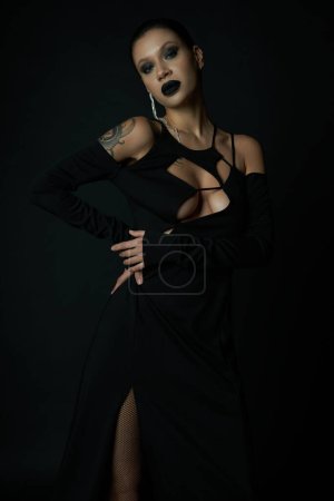 Tätowierte Frau in dunklem Make-up und sexy Halloween-Kleid im dunklen Studio vor der Kamera, schwarze Magie