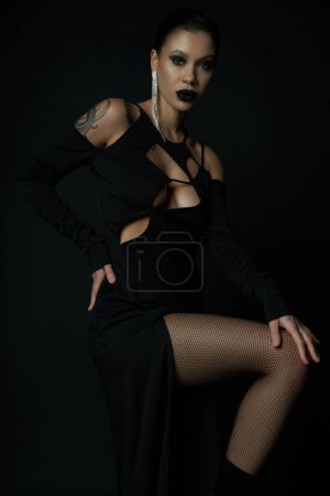 stylish tattooed woman in black halloween dress and creepy makeup posing with hand on hip on black
