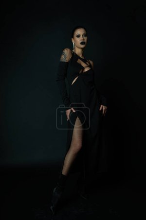 seductive tattooed woman in black halloween dress and dark makeup looking away while posing on black