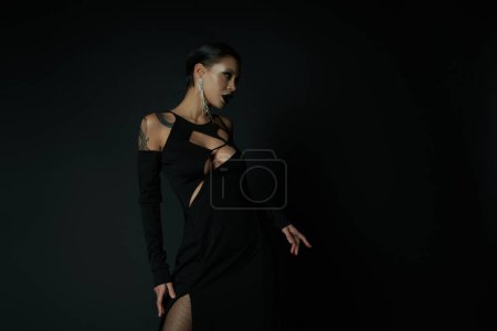 Foto de Sensualidad gótica, mujer tatuada en vestido de encantadora sexy y maquillaje oscuro mirando hacia otro lado en negro - Imagen libre de derechos