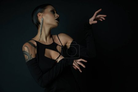 sexy tattooed woman in glamour halloween dress and creepy makeup gesturing on black backdrop