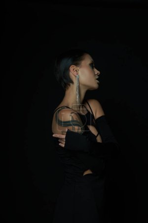 Photo for Side view of enchanting tattooed woman in black halloween dress and shiny earring on black backdrop - Royalty Free Image