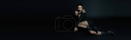 Photo for Sexy tattooed woman in black halloween dress sitting in darkness and looking at camera, banner - Royalty Free Image