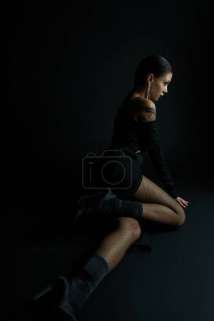 Téléchargez les photos : Femme enchanteresse en robe sexy et collants résille assis dans l'obscurité et regardant loin en studio - en image libre de droit