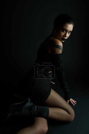 mujer apasionada en vestido sexy y medias de rejilla sentado y mirando a la cámara en el fondo negro