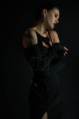 tattooed woman in stylish halloween dress and dark makeup posing with closed eyes on black backdrop