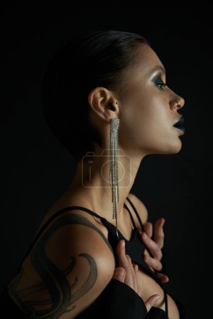 Photo for Profile of sensual tattooed woman with dark makeup and shiny earring on black, halloween concept - Royalty Free Image