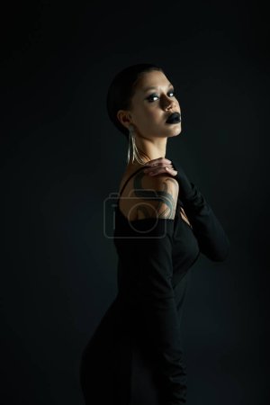 stylische tätowierte Frau mit dunklen Lippen trägt sexy Halloween-Kleid und schaut in die Kamera auf schwarz