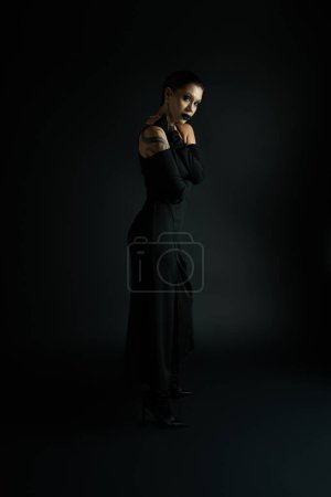 Foto de Sexy mujer tatuada con labios oscuros usando elegante vestido de halloween y mirando a la cámara en negro - Imagen libre de derechos